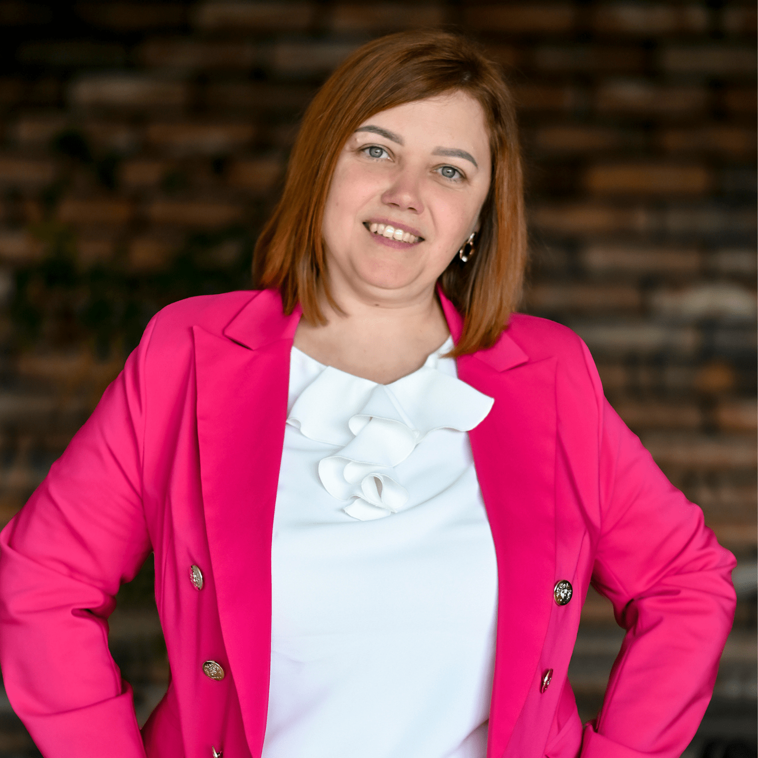 Kobieta w różowym żakiecie i białej bluzce z falbaną, uśmiechająca się w pozycji z rękami na biodrach, na tle ceramicznej ściany. Idealna jako przykład promocji usług, takich jak myjnie bezdotykowe.