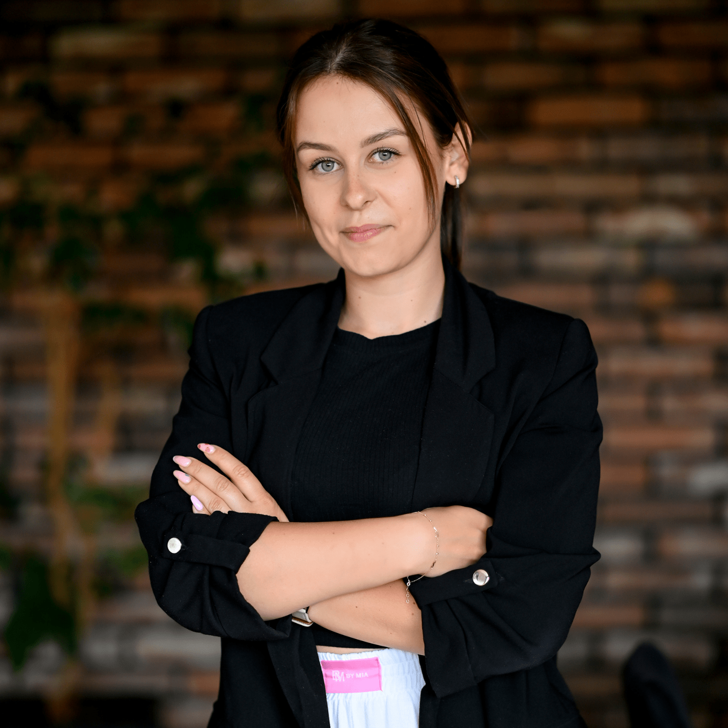 Młoda kobieta w czarnym żakiecie z założonymi rękami, uśmiechająca się, na tle ceglanej ściany. Wygląda pewnie i profesjonalnie, co może odzwierciedlać jej rolę w branży. Idealna dla producentów myjni bezdotykowych i marketingu w sektorze usług.