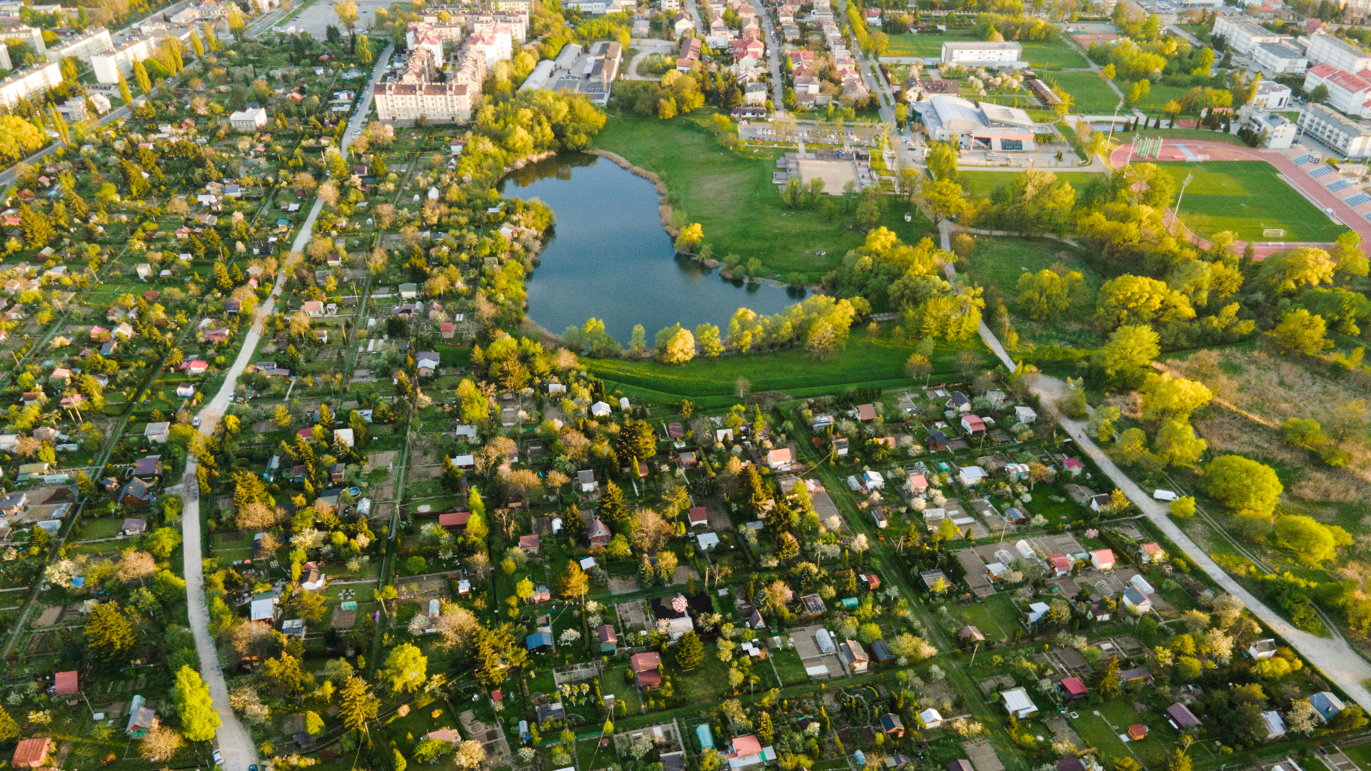 Widok z lotu ptaka na malowniczą okolicę z domami i ogrodami otaczającymi staw. W tle widać zieleń i inne zabudowania, tworząc idylliczny krajobraz. Idealne miejsce dla miłośników przyrody i relaksu wśród natury w kontekście myjni bezdotykowych dla samochodów.